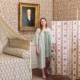 Une femme portant la XCOMM_NUISETTE CHARDON SAUGE se tient pieds nus dans une pièce vintage avec un papier peint floral, un lit assorti, un paravent et le portrait encadré d'un homme plus âgé derrière elle. la marque Toile de Jouy