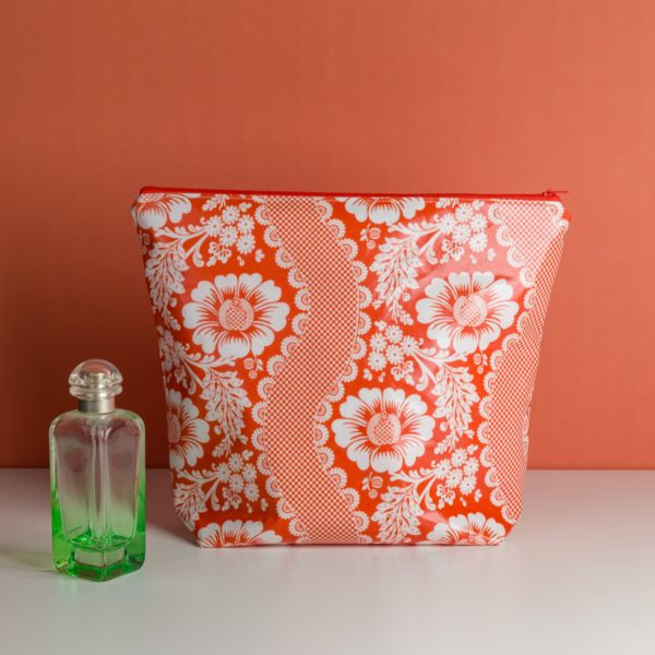 La « XCOMM_GRANDE TROUSSE DE TOILETTE FLEURS AMARANTE », une trousse de toilette à motifs floraux rouges et blancs, est posée à côté d'un flacon de parfum en verre vert muni d'un bouchon rond, sur un fond de couleur corail. la marque Toile de Jouy