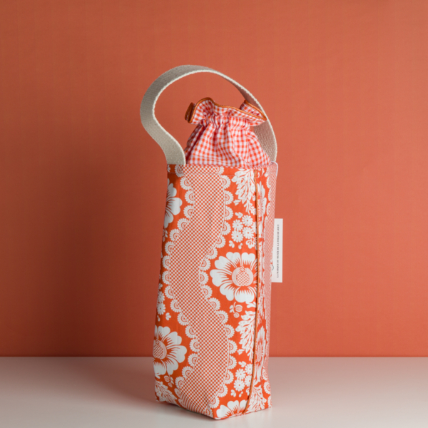 Le XCOMM_SAC ISOTHERME FLEURS MANDARINE est un sac en tissu à motif floral rouge et blanc avec une poignée crème et un cordon vichy rouge, présenté à la verticale sur un fond rouge assorti. la marque Toile de Jouy