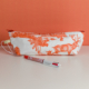 La trousse XCOMM_TROUSSE POULETTE4 PARTIES MANDARINE, une trousse en tissu blanc et orange au motif inspiré de la nature, repose sur une surface blanche, avec un stylo rouge et blanc placé devant, sur fond orange. la marque Toile de Jouy