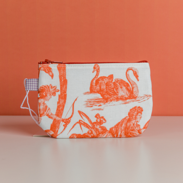 Le modèle XCOMM_TROUSSE POULETTE4 PARTIES MANDARINE est une petite pochette en tissu dotée d'une fermeture éclair rouge, ornée d'illustrations représentant des cygnes et des personnages de couleur orange sur fond blanc, représentés sur une surface blanche devant un mur orange. la marque Toile de Jouy