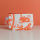 Le modèle XCOMM_TROUSSE POULETTE4 PARTIES MANDARINE est une petite pochette en tissu dotée d'une fermeture éclair rouge, ornée d'illustrations représentant des cygnes et des personnages de couleur orange sur fond blanc, représentés sur une surface blanche devant un mur orange. la marque Toile de Jouy