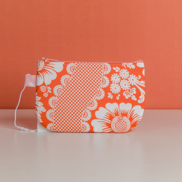 La XCOMM_TROUSSE STYLOS FLEURS MANDARINE est une petite pochette à fermeture éclair avec un motif floral et à carreaux orange et blanc, comportant une étiquette en tissu rose avec des étoiles blanches, présentée sur une surface blanche sur fond orange. la marque Toile de Jouy