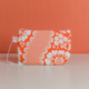 La XCOMM_TROUSSE STYLOS FLEURS MANDARINE est une petite pochette à fermeture éclair avec un motif floral et à carreaux orange et blanc, comportant une étiquette en tissu rose avec des étoiles blanches, présentée sur une surface blanche sur fond orange. la marque Toile de Jouy
