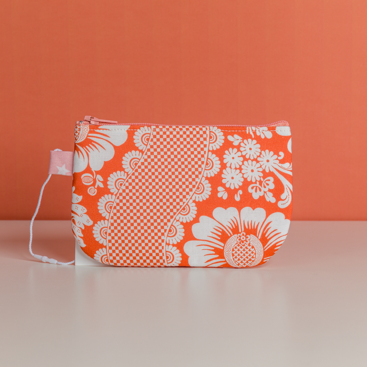 La XCOMM_TROUSSE STYLOS FLEURS MANDARINE est une petite pochette à fermeture éclair avec un motif floral et à carreaux orange et blanc, comportant une étiquette en tissu rose avec des étoiles blanches, présentée sur une surface blanche sur fond orange. la marque Toile de Jouy