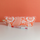 La XCOMM_TROUSSE STYLOS FLEURS MANDARINE, une trousse à crayons orange et blanche à motifs floraux, repose sur une surface blanche avec un stylo rouge et blanc devant. Le fond est orange uni. la marque Toile de Jouy