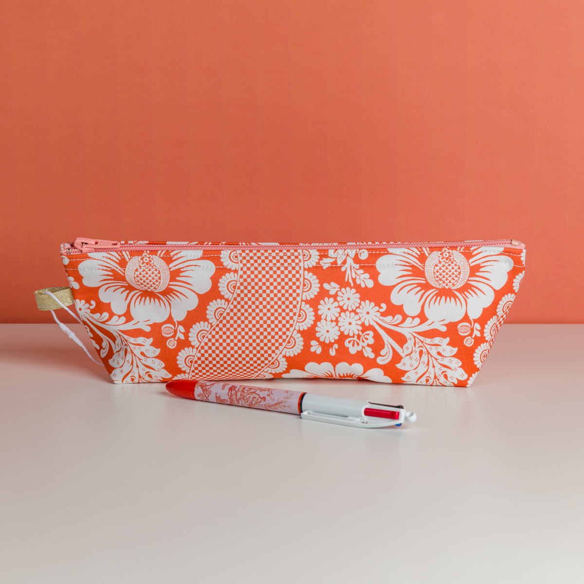 La XCOMM_TROUSSE STYLOS FLEURS MANDARINE, une trousse à crayons orange et blanche à motifs floraux, repose sur une surface blanche avec un stylo rouge et blanc devant. Le fond est orange uni. la marque Toile de Jouy