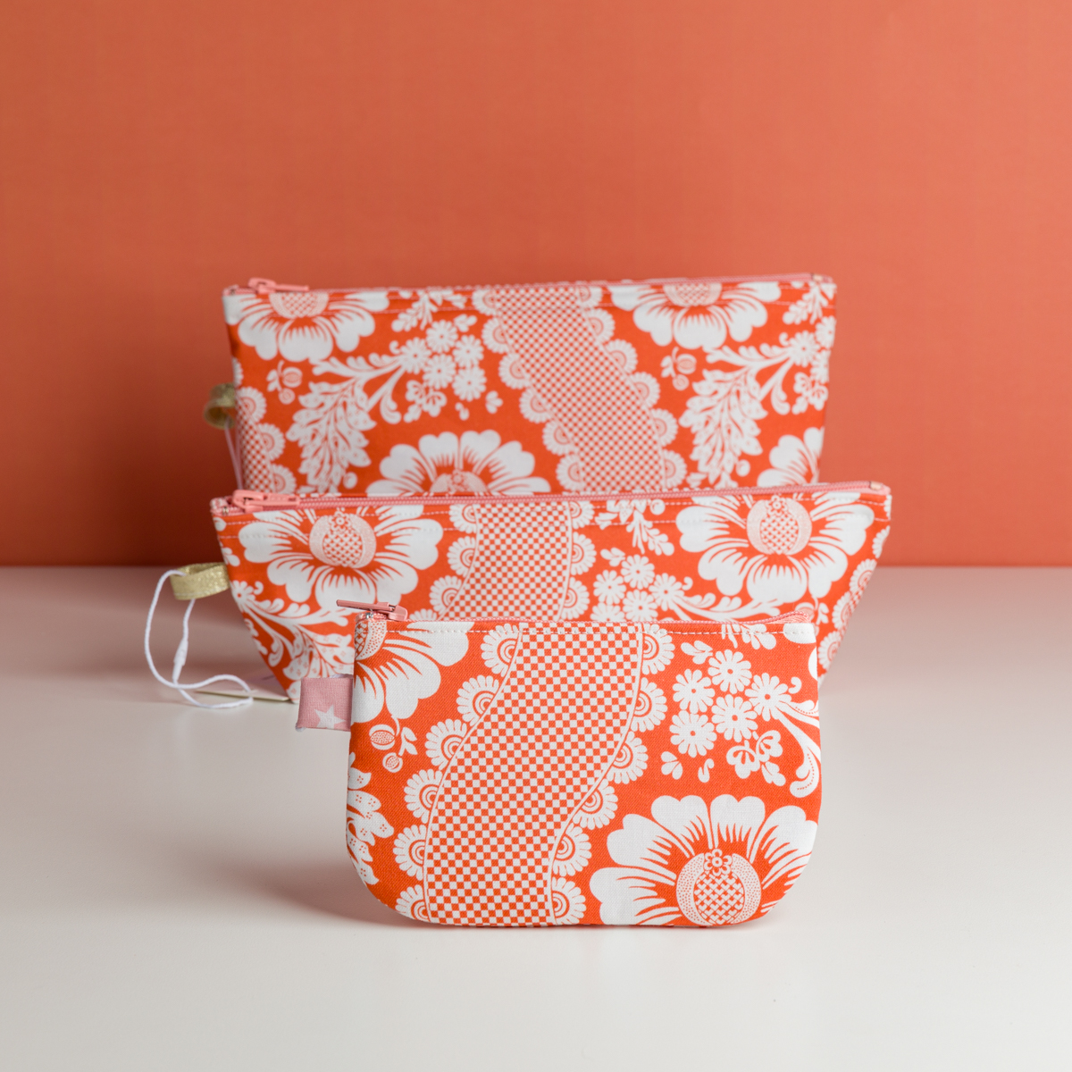 Trois pochettes à fermeture éclair XCOMM_TROUSSE STYLOS FLEURS MANDARINE avec des motifs floraux rouges et blancs sont disposées par taille, du plus petit au plus grand, sur une surface blanche, sur un fond rouge assorti. la marque Toile de Jouy