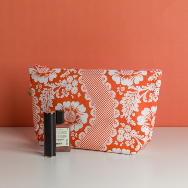 La XCOMM_TROUSSE STYLOS FLEURS MANDARINE, une trousse de maquillage orange et blanche à motifs floraux, est présentée sur une surface blanche à fond corail à côté d'un tube de rouge à lèvres noir et d'un vernis à ongles rose. la marque Toile de Jouy
