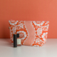 La XCOMM_TROUSSE STYLOS FLEURS MANDARINE, une trousse de maquillage orange et blanche à motifs floraux, est présentée sur une surface blanche à fond corail à côté d'un tube de rouge à lèvres noir et d'un vernis à ongles rose. la marque Toile de Jouy
