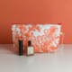 La pochette XCOMM_TROUSSE POULETTE4 PARTIES MANDARINE, une pochette à fermeture éclair ornée d'un motif « toile » orange et blanc, repose sur une surface blanche, devant laquelle se trouvent un tube de rouge à lèvres noir et un flacon de parfum carré, le tout sur fond orange. la marque Toile de Jouy
