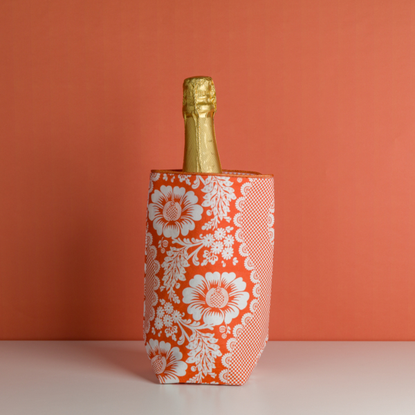 Le XCOMM_HOUSSE BOUTEILLE FLEURS MANDARINE, un sac isotherme à motifs floraux rouges et blancs, contient une bouteille de champagne enveloppée d'une feuille d'or sur une surface blanche avec un arrière-plan orange. la marque Toile de Jouy