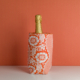 Le XCOMM_HOUSSE BOUTEILLE FLEURS MANDARINE, un sac isotherme à motifs floraux rouges et blancs, contient une bouteille de champagne enveloppée d'une feuille d'or sur une surface blanche avec un arrière-plan orange. la marque Toile de Jouy