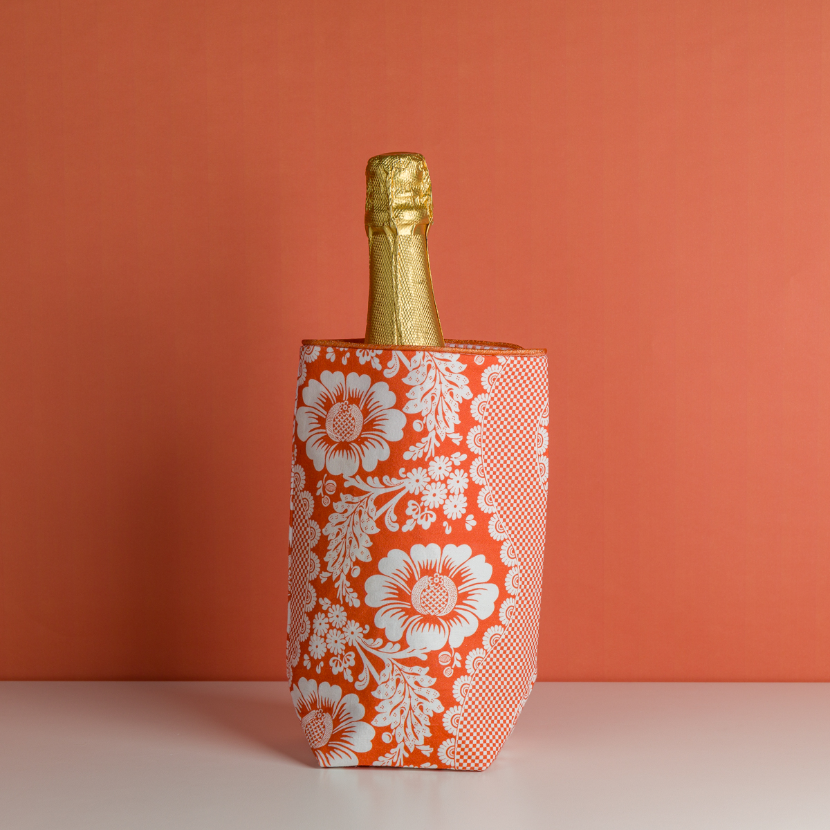 Le XCOMM_HOUSSE BOUTEILLE FLEURS MANDARINE, un sac isotherme à motifs floraux rouges et blancs, contient une bouteille de champagne enveloppée d'une feuille d'or sur une surface blanche avec un arrière-plan orange. la marque Toile de Jouy