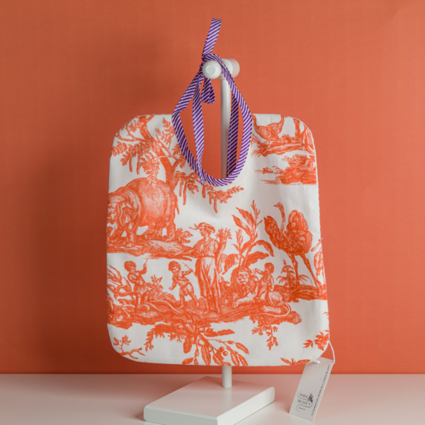 Un bavoir pour bébé XCOMM_BAVOIR 4 PARTIES MANDARINE, orné d'un motif blanc et orange et d'une cravate à rayures violettes, présenté sur un présentoir devant un fond orange, avec une étiquette produit suspendue à l'un des coins. la marque Toile de Jouy