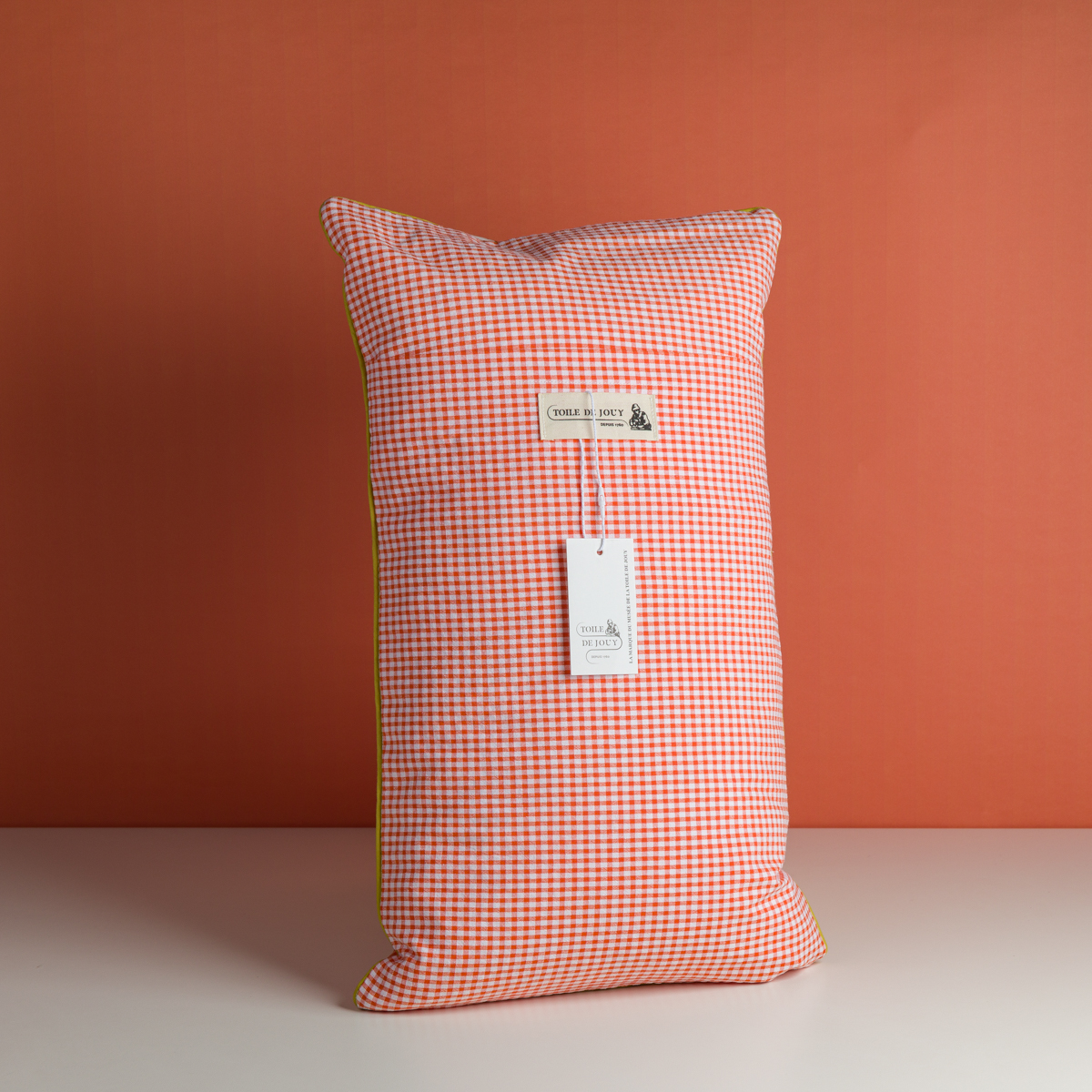 Le XCOMM_COUSSIN 30X50 FLEURS MANDARINE, un oreiller rectangulaire, se tient debout sur un fond orange. Une étiquette blanche avec du texte et une petite illustration est attachée au centre de l'oreiller. la marque Toile de Jouy