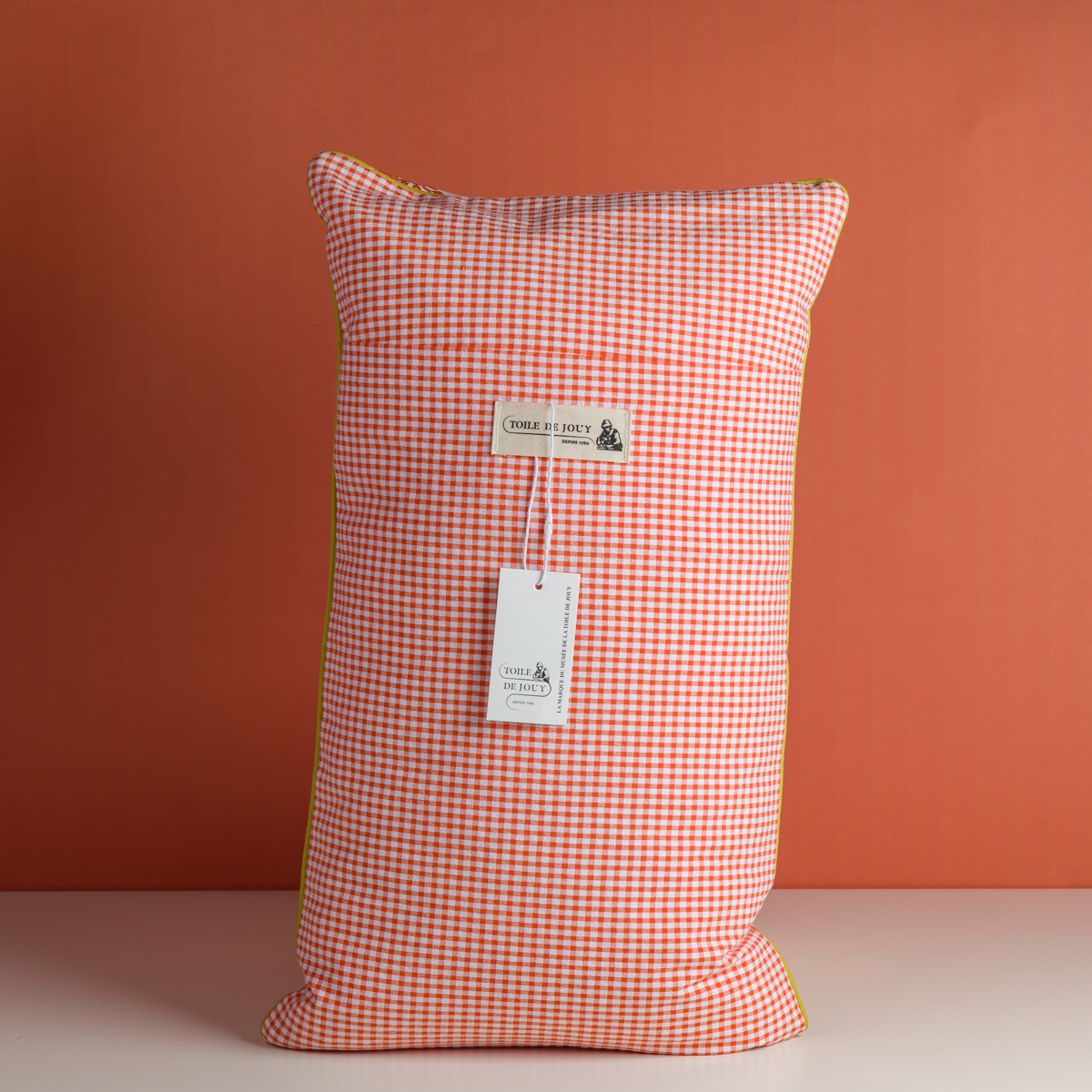 Un oreiller rectangulaire, "XCOMM_COUSSIN 30X50 FLEURS MANDARINE", avec un motif floral se tient debout sur un fond orange. Une étiquette portant la mention "COOKIE & PONEY" est attachée à l'avant de l'oreiller. la marque Toile de Jouy