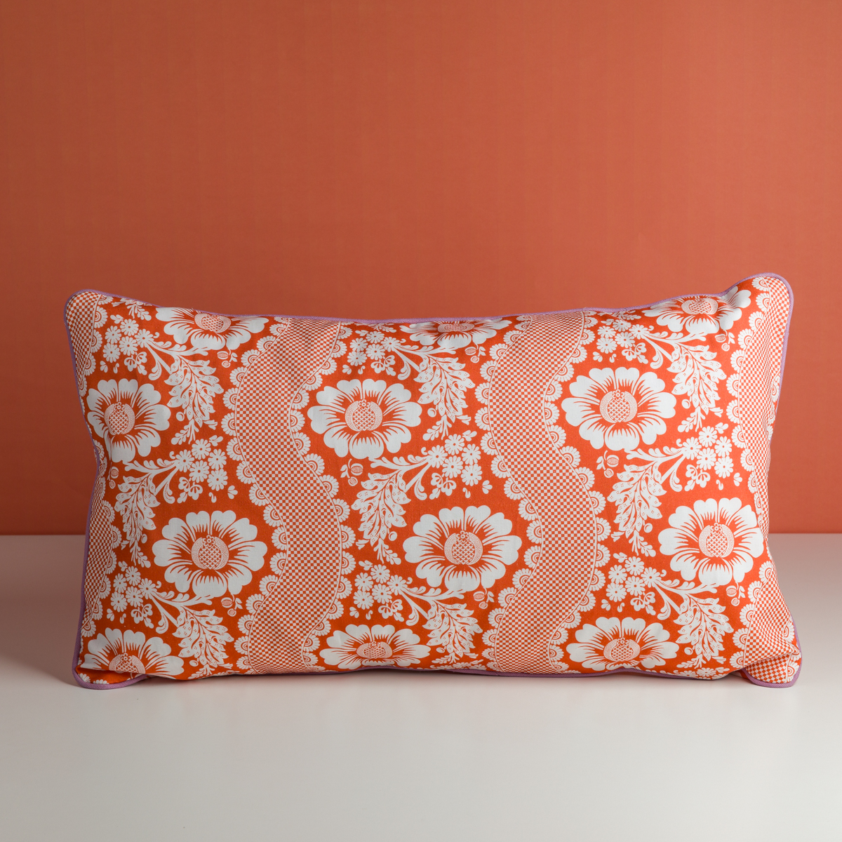 Le XCOMM_COUSSIN 30X50 FLEURS MANDARINE, un coussin rectangulaire avec un motif floral rouge et blanc composé de grandes fleurs et de rayures en forme de dentelle, repose sur une surface blanche sur un fond rouge assorti. la marque Toile de Jouy