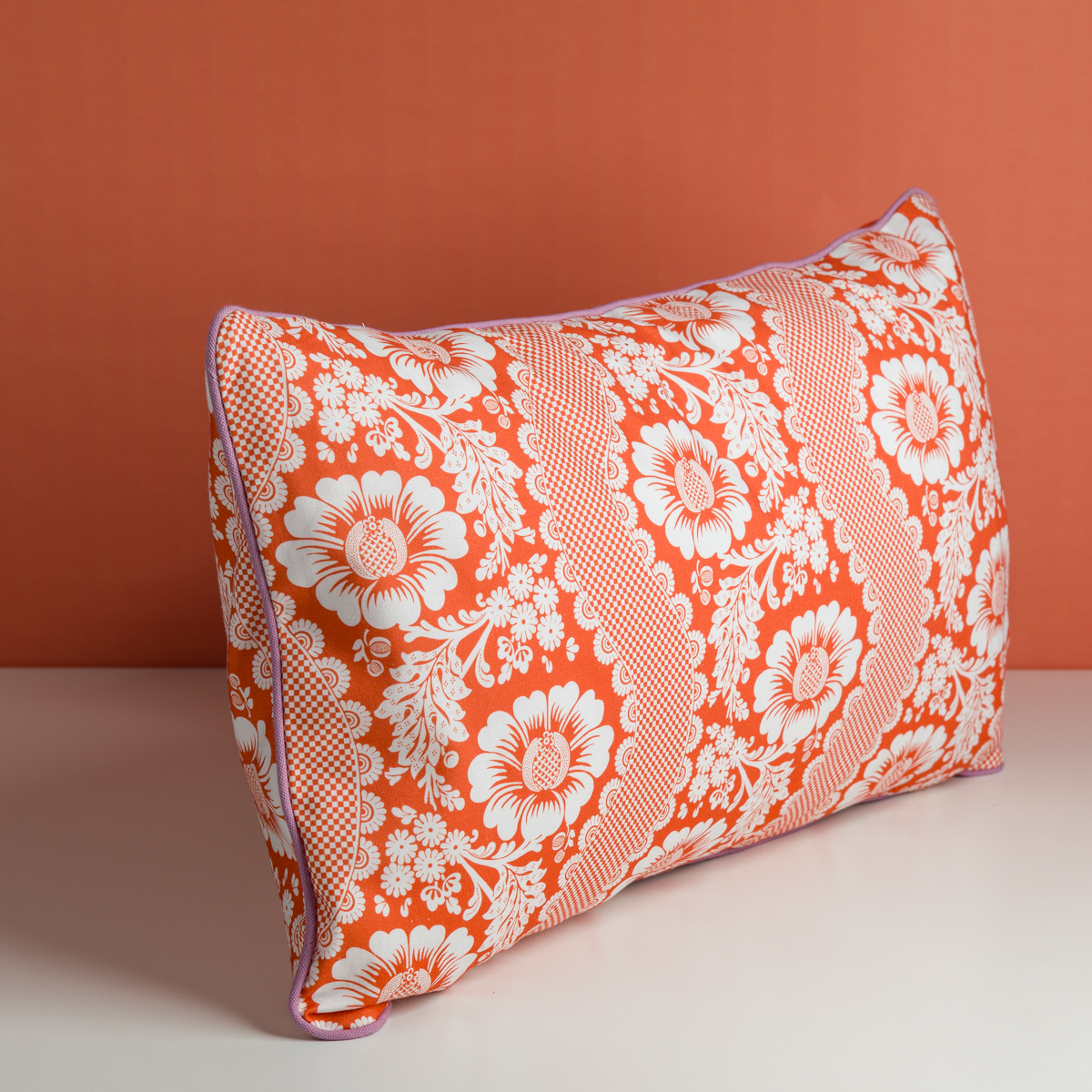 Le XCOMM_COUSSIN 30X50 FLEURS MANDARINE, un coussin rectangulaire avec des motifs floraux blancs sur un fond orange vif, est placé sur une surface blanche devant un mur orange. la marque Toile de Jouy
