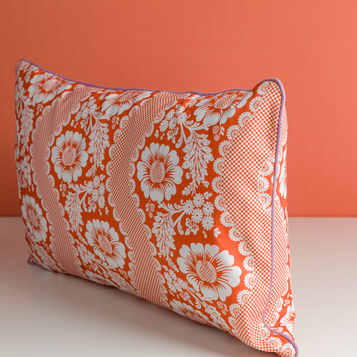 Le coussin décoratif XCOMM_COUSSIN 30X50 FLEURS MANDARINE présente un motif floral rouge et blanc avec un liseré violet, reposant sur une surface blanche sur un fond rouge assorti. la marque Toile de Jouy