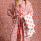 Une femme se tient pieds nus sur un sol à motifs, portant le XCOMM_KIMONO EPIS MANDARINE - une robe à motifs roses et blancs - et tenant un tissu à motifs rouges. L'arrière-plan coordonné est assorti à sa tenue, créant ainsi une scène à motifs unifiée. la marque Toile de Jouy