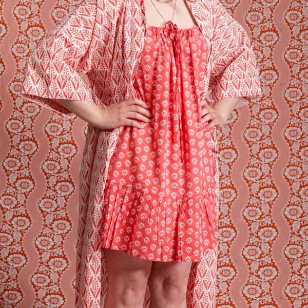 Une femme debout, pieds nus, mains sur les hanches, portant le XCOMM_KIMONO EPIS MANDARINE - une robe rose à motifs et un peignoir assorti - contre un mur et un sol aux motifs géométriques roses similaires, se fondant dans le décor pour un effet de camouflage. la marque Toile de Jouy
