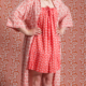 Une femme debout, pieds nus, mains sur les hanches, portant le XCOMM_KIMONO EPIS MANDARINE - une robe rose à motifs et un peignoir assorti - contre un mur et un sol aux motifs géométriques roses similaires, se fondant dans le décor pour un effet de camouflage. la marque Toile de Jouy