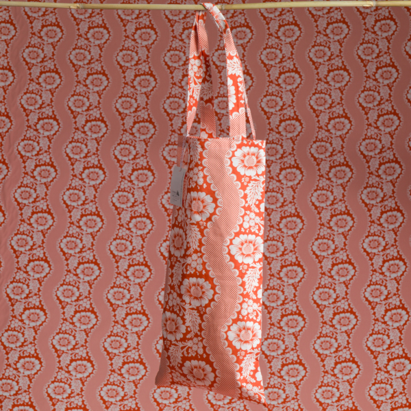 Le sac fourre-tout XCOMM_SAC ISOTHERME FLEURS MANDARINE, aux motifs floraux et ondulés orange et blancs éclatants, est suspendu à une tige en bois sur un fond à motifs assortis. la marque Toile de Jouy