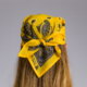 On voit de dos une personne aux longs cheveux châtain clair portant le XCOMM_BANDANA BOTEH JAINE 50X50, un bandana jaune à motifs paisley noué à l'arrière de la tête, sur un fond gris uni. la marque Toile de Jouy