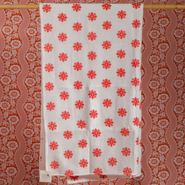 La XCOMM_SERVIETTE HAMMAM EPIS MANDARINE 100X180, une serviette blanche avec un motif floral rouge et une petite étiquette blanche, est suspendue à une tringle dorée contre un papier peint rétro ondulé à motifs floraux roses et rouges. la marque Toile de Jouy