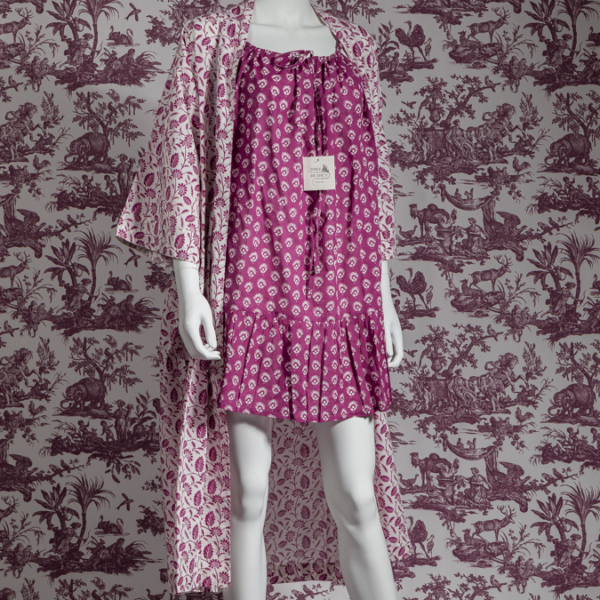 Un mannequin présente la XCOMM_NUISETTE CHARDON AMARANTE - une robe magenta courte et sans manches avec un imprimé floral blanc et une robe longue assortie - sur un papier peint en toile violette et blanche. la marque Toile de Jouy