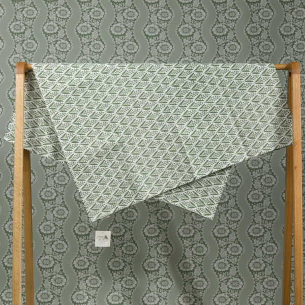 Le XCOMM_SERVIETTE HAMMAM EPIS MANDARINE 100X180, au motif vert et blanc, est drapé sur un support en bois devant un papier peint assorti pour un effet camouflé. Une petite étiquette blanche est accrochée au tissu. la marque Toile de Jouy
