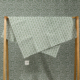 Le XCOMM_SERVIETTE HAMMAM EPIS MANDARINE 100X180, au motif vert et blanc, est drapé sur un support en bois devant un papier peint assorti pour un effet camouflé. Une petite étiquette blanche est accrochée au tissu. la marque Toile de Jouy