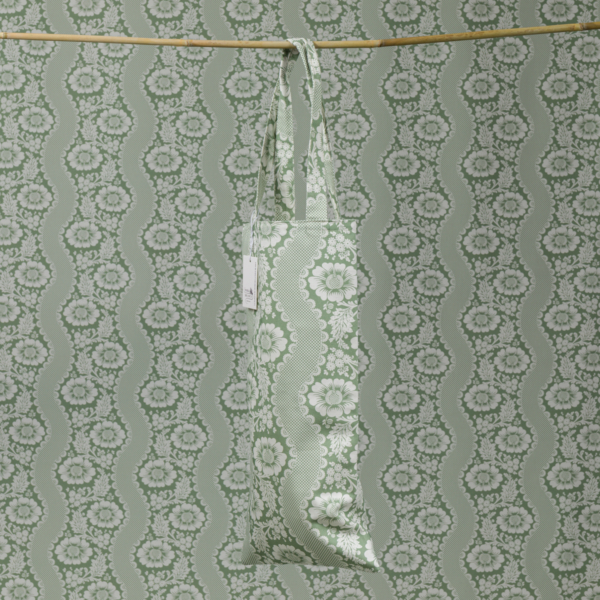 Le XCOMM_HOUSSE BOUTEILLE OFFRANDE SAUGE, avec son motif vert et blanc, est suspendu à une tringle en bois et s'intègre parfaitement dans un fond de papier peint à motifs floraux et ondulés assorti. la marque Toile de Jouy