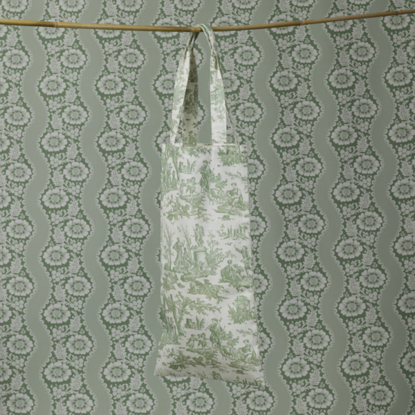 Le XCOMM_HOUSSE BOUTEILLE OFFRANDE SAUGE, aux motifs pastoraux verts, est suspendu à une tringle et s'intègre parfaitement à un fond de papier peint floral ondulé vert et blanc coordonné. la marque Toile de Jouy