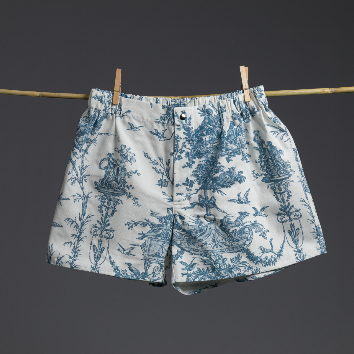 Le XCOMM_COUSSIN 30X50 ESCARPOLETTE BLEU, orné d'un motif de toile bleue, est suspendu à une tige de bambou à l'aide de deux pinces à linge en bois, sur un fond uni foncé. la marque Toile de Jouy