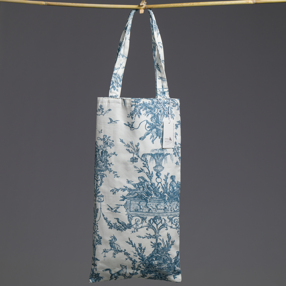 Un sac fourre-tout blanc, baptisé « XCOMM_COUSSIN 30X50 ESCARPOLETTE BLEU », orné de motifs floraux et d'oiseaux bleus très détaillés, est suspendu à une tringle horizontale par ses deux anses. Une petite étiquette blanche est fixée au sac, sur un fond gris uni. la marque Toile de Jouy