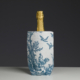 Une bouteille de champagne ornée d'une capsule en feuille d'or est placée dans la housse en tissu XCOMM_COUSSIN 30X50 ESCARPOLETTE BLEU, qui représente une scène champêtre bleue et blanche avec des oiseaux et un coq, sur fond sombre. la marque Toile de Jouy