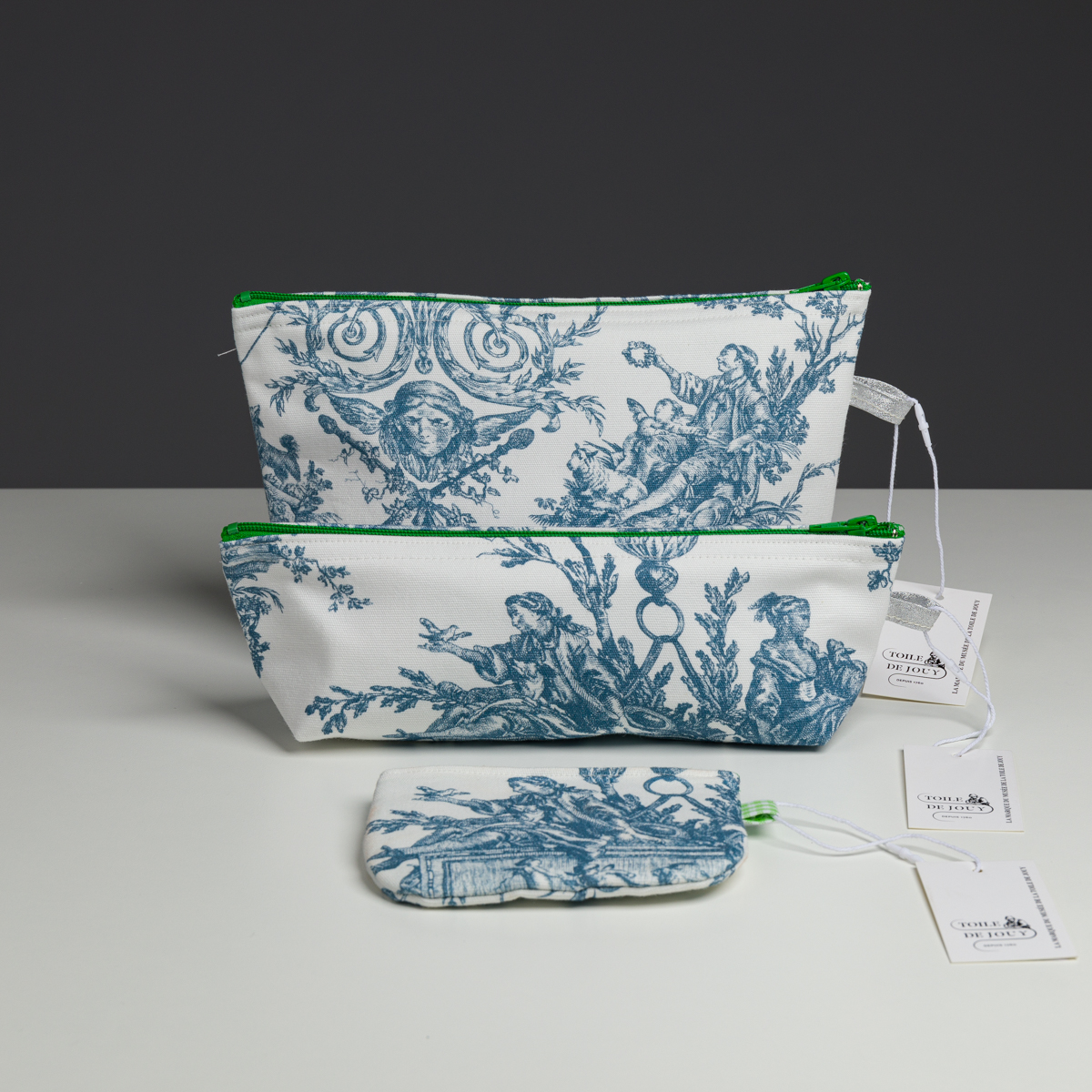 Trois pochettes en tissu blanc, ornées de motifs « Escarpolette » bleus sur toile et dotées de fermetures éclair vertes, sont disposées sur une surface blanche. Chaque pochette, issue de la collection XCOMM_COUSSIN 30X50 ESCARPOLETTE BLEU, est munie d’une étiquette fixée sur le côté droit et se décline en différentes tailles, de la plus grande à la plus petite. la marque Toile de Jouy