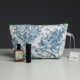 Un sac XCOMM_COUSSIN 30X50 ESCARPOLETTE BLEU, muni d'une fermeture éclair verte, est posé sur une table. Devant lui se trouvent une petite bouteille marron et un récipient cylindrique noir. Une étiquette blanche est accrochée à la poignée latérale du sac. la marque Toile de Jouy