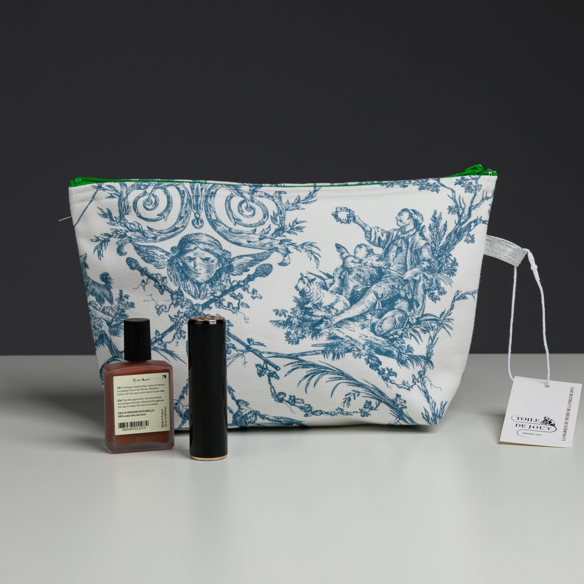 Un sac XCOMM_COUSSIN 30X50 ESCARPOLETTE BLEU, muni d'une fermeture éclair verte, est posé sur une table. Devant lui se trouvent une petite bouteille marron et un récipient cylindrique noir. Une étiquette blanche est accrochée à la poignée latérale du sac. la marque Toile de Jouy