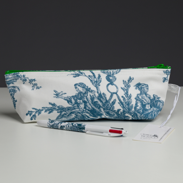 XCOMM_COUSSIN 30X50 ESCARPOLETTE BLEU, orné d'un tissu à motifs blancs et bleus et doté d'une fermeture éclair verte, se détache sur un fond sombre. À l'avant de la pochette se trouvent un stylo multicolore et une étiquette. la marque Toile de Jouy