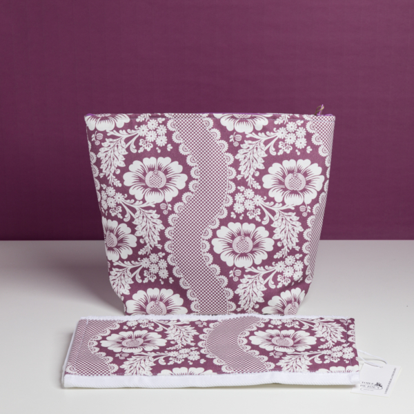 La « XCOMM_GRANDE TROUSSE DE TOILETTE FLEURS AMARANTE », composée d'une pochette à fermeture éclair et d'un accessoire en tissu assorti orné d'un motif de dentelle florale violette et blanche, est présentée sur un fond violet avec une petite étiquette attachée. la marque Toile de Jouy