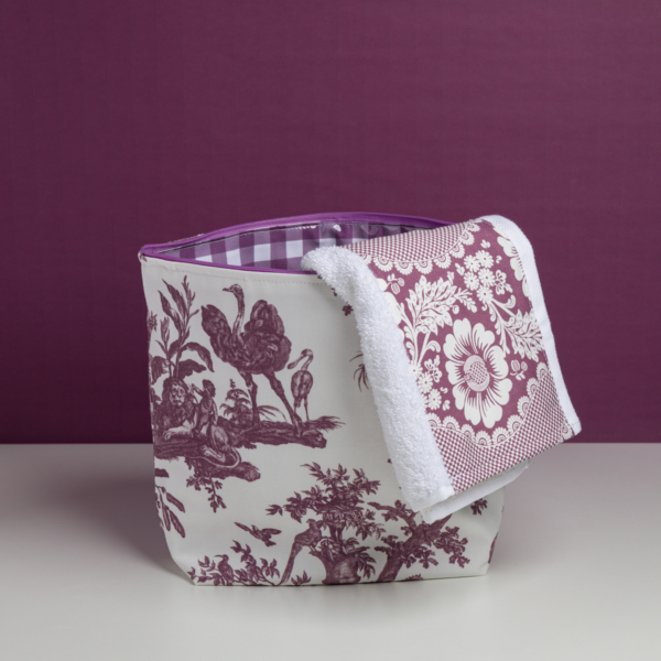 La table XCOMM_TROUSSE POULETTE4 PARTIES MANDARINE, ornée d'un motif « toile » champêtre violet et blanc, repose sur un plateau blanc devant un fond violet, avec une serviette assortie drapée sur son bord. la marque Toile de Jouy