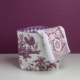 La table XCOMM_TROUSSE POULETTE4 PARTIES MANDARINE, ornée d'un motif « toile » champêtre violet et blanc, repose sur un plateau blanc devant un fond violet, avec une serviette assortie drapée sur son bord. la marque Toile de Jouy
