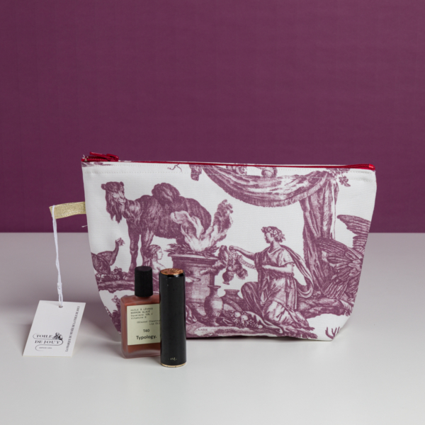 La trousse XCOMM_TROUSSE POULETTE4 PARTIES MANDARINE, une trousse de maquillage blanche ornée d'un imprimé champêtre violet, repose sur une surface blanche, accompagnée de son étiquette, d'un flacon de parfum et d'un rouge à lèvres noir. L'arrière-plan est violet. la marque Toile de Jouy