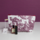 La trousse XCOMM_TROUSSE POULETTE4 PARTIES MANDARINE, une trousse de maquillage blanche ornée d'un imprimé champêtre violet, repose sur une surface blanche, accompagnée de son étiquette, d'un flacon de parfum et d'un rouge à lèvres noir. L'arrière-plan est violet. la marque Toile de Jouy