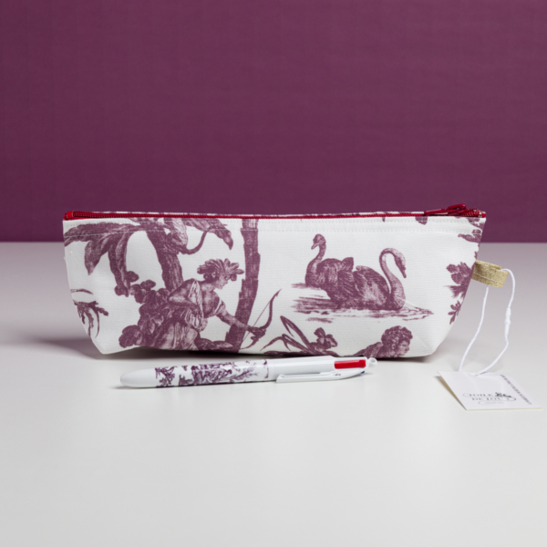 La trousse XCOMM_TROUSSE POULETTE4 PARTIES MANDARINE, une trousse blanche ornée d'un motif de cygne et d'arbres sur toile violette, est présentée sur une surface blanche, avec un stylo et une étiquette assortis placés devant, sur fond violet. la marque Toile de Jouy