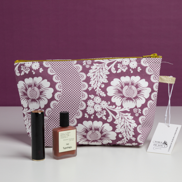 La XCOMM_TROUSSE STYLOS FLEURS AMARANTE, une trousse de maquillage à fleurs violettes et blanches avec une étiquette, est posée sur une surface blanche avec un rouge à lèvres et un flacon de typologie devant, sur un fond violet assorti. la marque Toile de Jouy