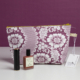 La XCOMM_TROUSSE STYLOS FLEURS AMARANTE, une trousse de maquillage à fleurs violettes et blanches avec une étiquette, est posée sur une surface blanche avec un rouge à lèvres et un flacon de typologie devant, sur un fond violet assorti. la marque Toile de Jouy